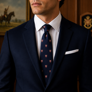 A man wearing a navy blue suit jacket, white dress shirt, and a navy blue patterned tie with a white pocket square, photographed in an indoor setting with wood paneling and framed artwork in the background.